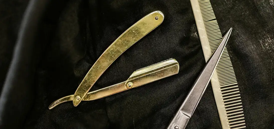Close-up of barber tools: straight razor, scissors, and comb on dark fabric