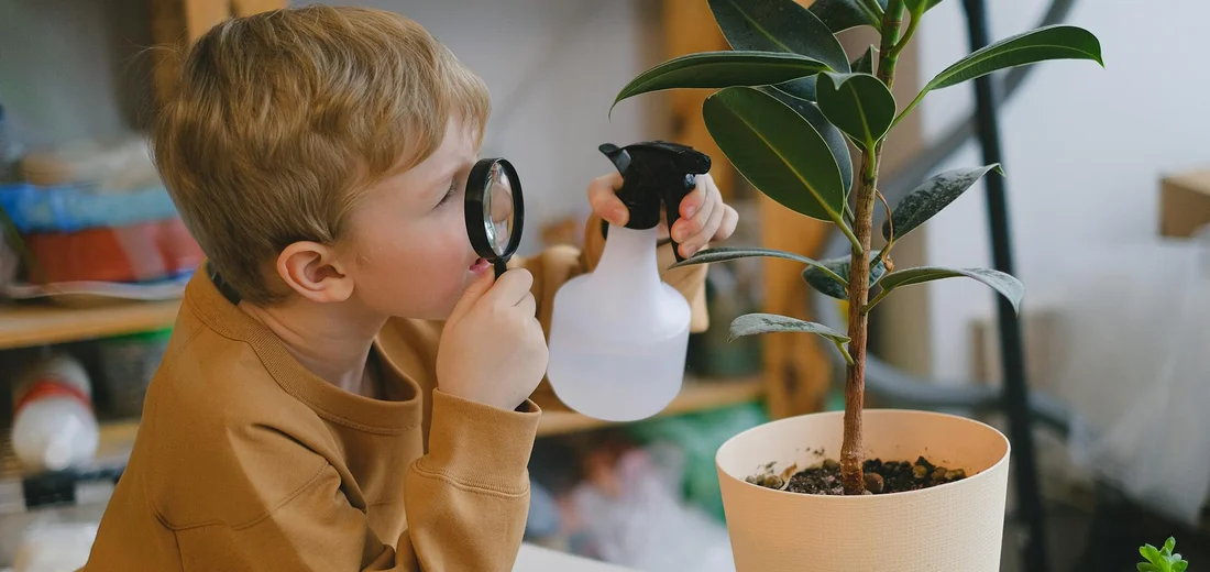 A young child uses a spray bottle and a magnifying glass to inspect a potted plant.