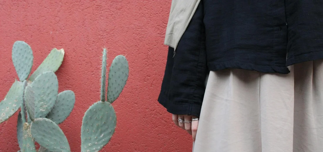Person in a dark jacket and beige skirt standing beside a cactus against a red wall.