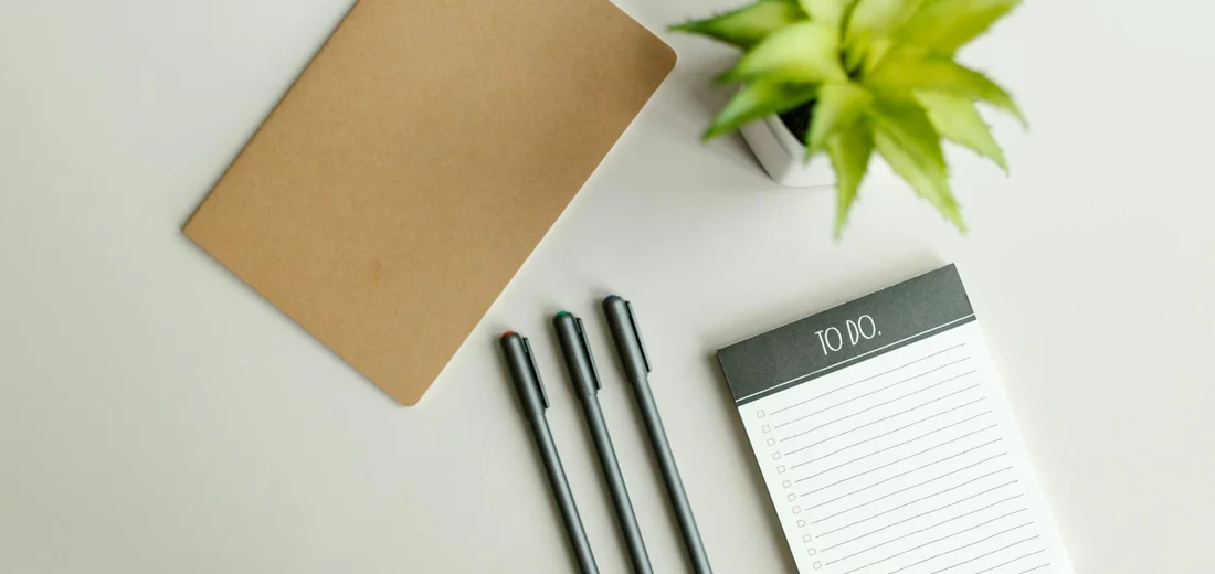 Desk setup featuring a small potted succulent, a brown notebook, three black pens, and a to-do list pad, suggesting planning for quarantine and treatment of succulents.