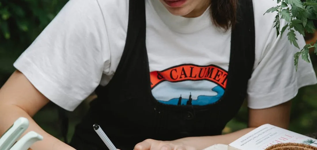 Close-up of a person wearing a white t-shirt and black apron, writing in a notebook beside an open book, with greenery in the background, illustrating planning to amend soil for succulents.