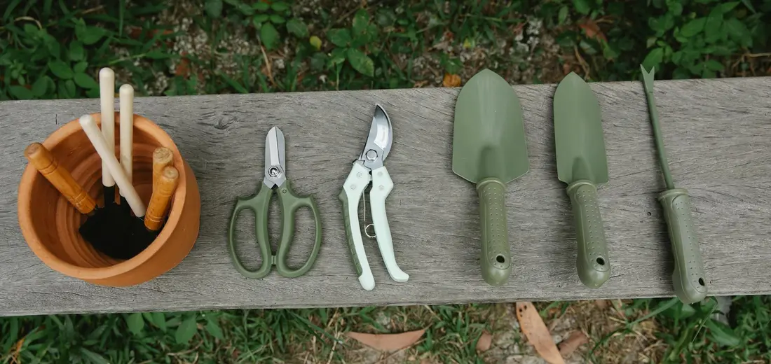 Garden tools laid out on a wooden surface: a terracotta pot with wooden stakes, pruning scissors, pruning shears, and three green-handled trowels.