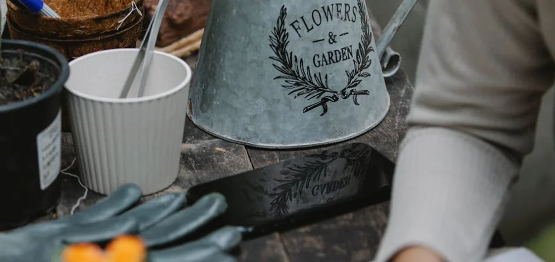 Close-up of gardening tools on a work table, including a metal watering can labeled 'Flowers & Garden', a white cup, and a pair of blue gloves.