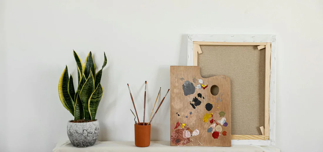 Potted snake plant on a white table with a small orange pot holding painting brushes, a wooden artist's palette with paint, and a blank frame leaning against the wall.