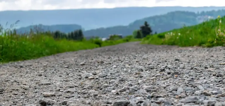 Close-up of a gravel path with small rocks, green vegetation on the sides, and distant hills.