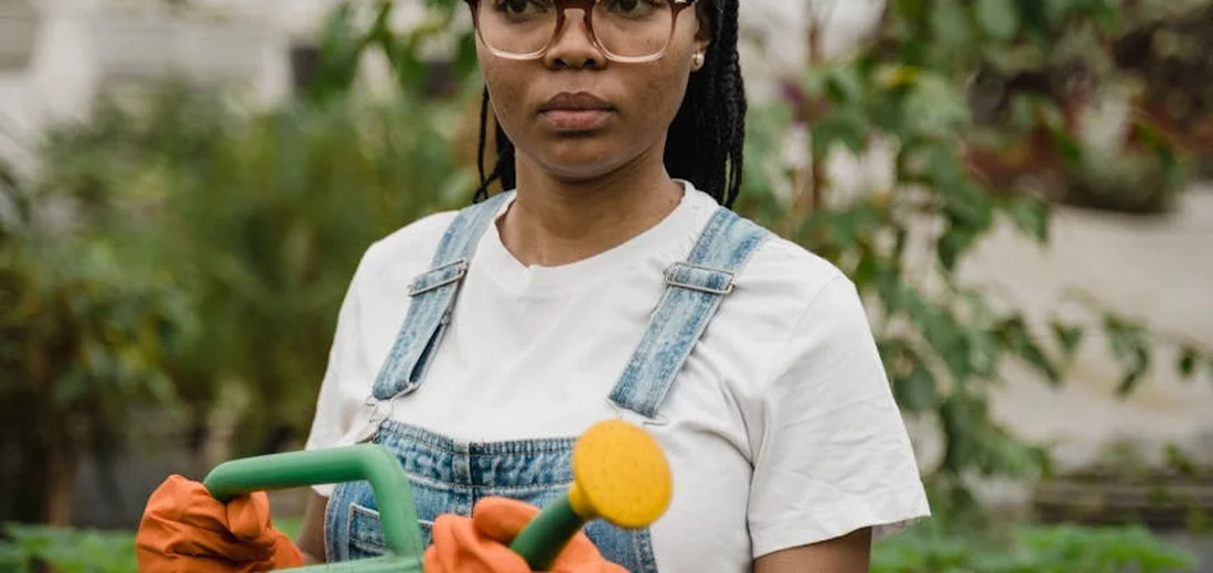 Gardener wearing a white shirt and denim overalls with orange gloves holds a green-handled garden tool in a lush outdoor garden