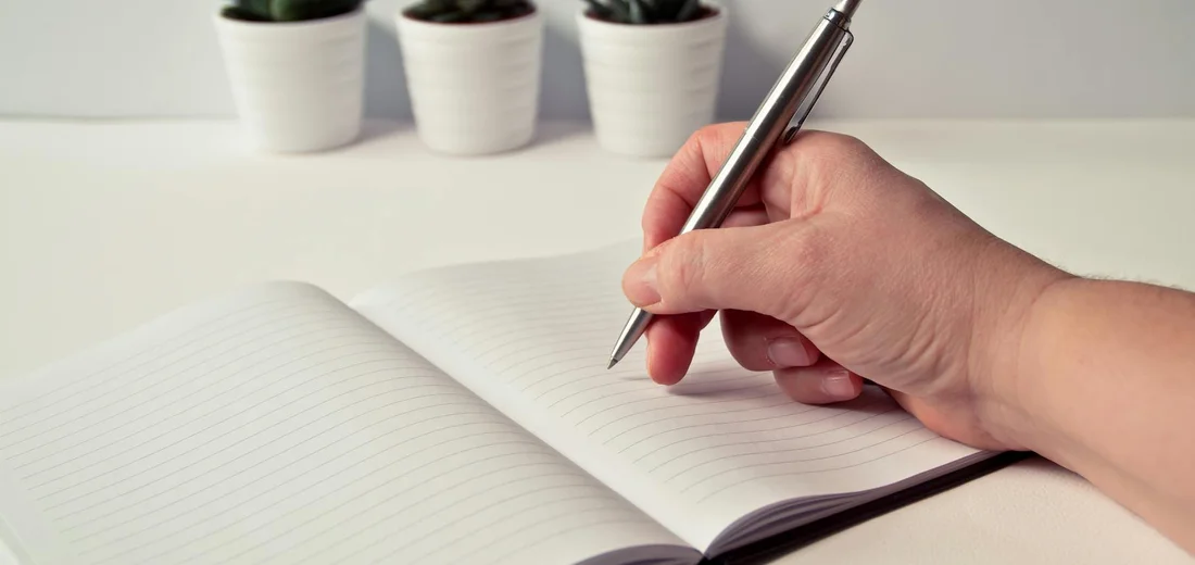 Close-up of a hand holding a pen over an open notebook, with small potted succulents blurred in the background.