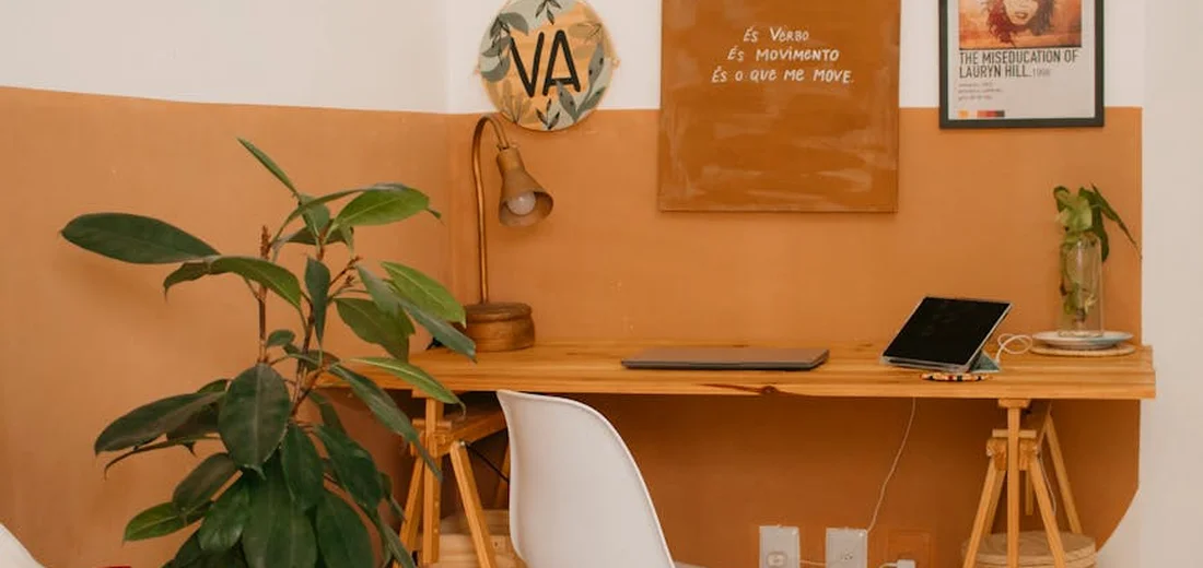 A home office corner with a wooden desk, a large leafy houseplant on the left, peach walls, framed art, and a laptop on the desk.