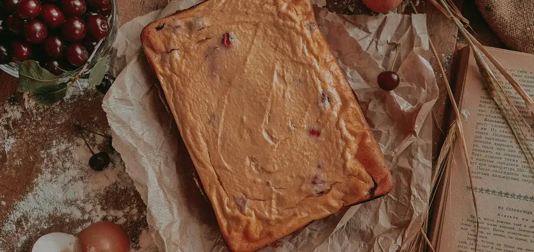 A rectangular loaf cake sits on parchment on a rustic surface, with berries and baking ingredients around it.