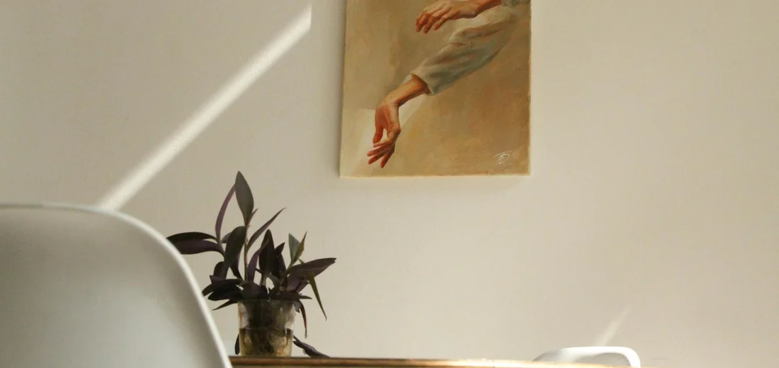 Potted plant on a wooden table beside a white chair, with a painting of an arm reaching downward on the wall.