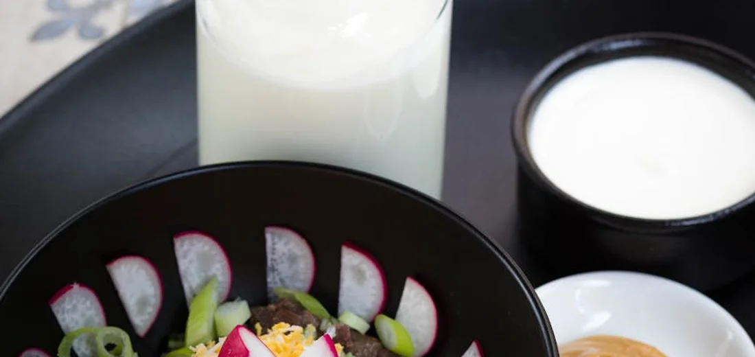 Glass pitcher of milk and bowls of dairy products on a dark surface beside a colorful salad.