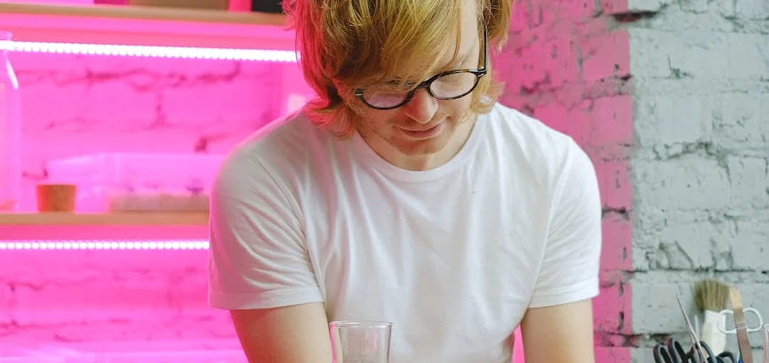Person in a white t-shirt and glasses stands at a table in a bright room with pink neon lighting and a white brick wall.