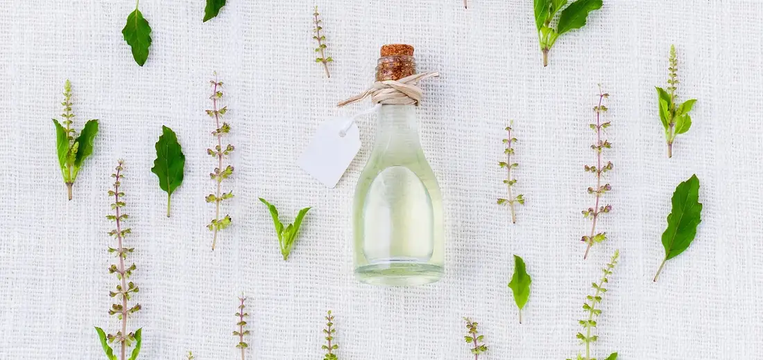 Glass bottle with a cork and tag surrounded by green leaves and stems on a light textured surface