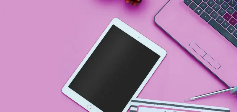 Flat lay of a pink workspace featuring a tablet, a laptop, and a pen on a pink surface.
