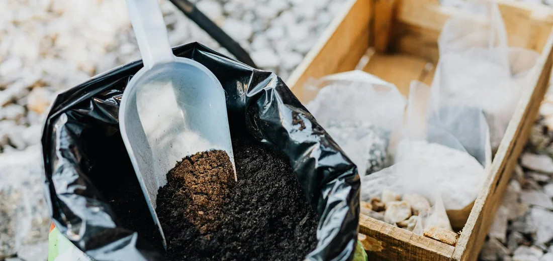A scoop of dark potting soil rests on an open bag, with small bags of amendments in a wooden crate beside it.