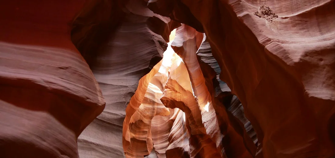 Sunlight streams through a narrow slot canyon, illuminating smooth red rock walls.