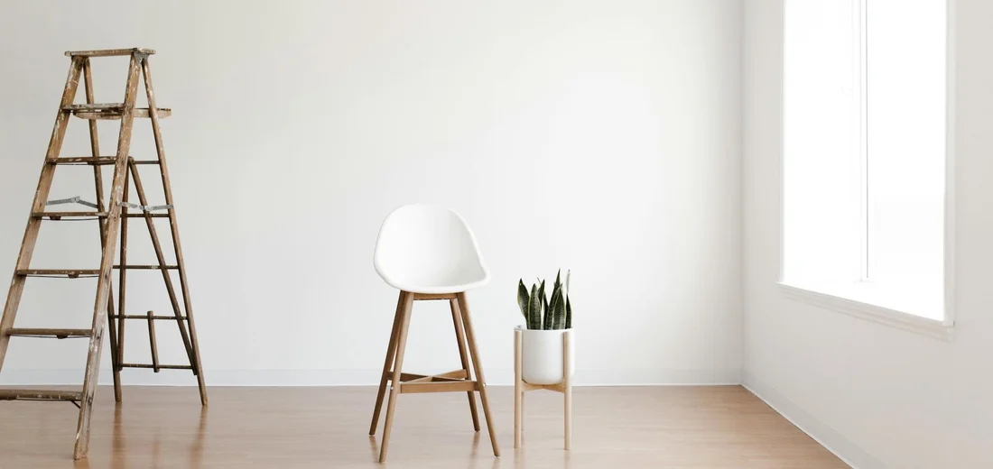 Minimalist interior with a wooden ladder on the left, a white chair in the center, and a small snake plant in a light ceramic pot on a wooden floor near a window.