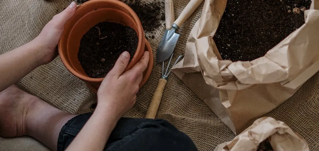 Close-up of a person holding a small terracotta pot with soil, surrounded by gardening tools and brown paper bags on a burlap surface.