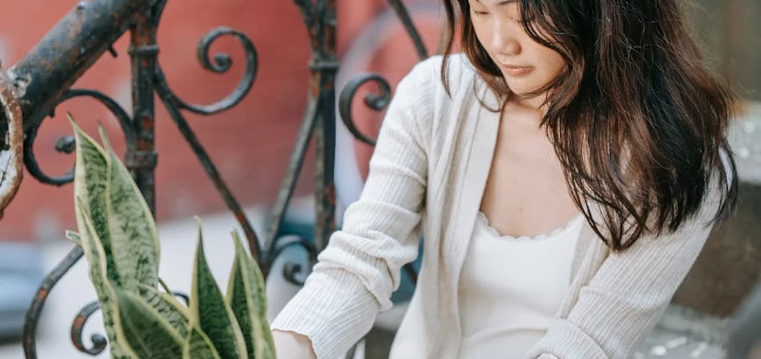 A woman with long dark hair wearing a white cardigan sits beside a snake plant on a decorative balcony, demonstrating gentle, patient care.