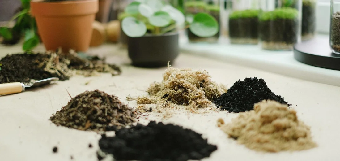 Multiple piles of soil and compost arranged on a work surface with a trowel, and potted plants on a windowsill in the background.