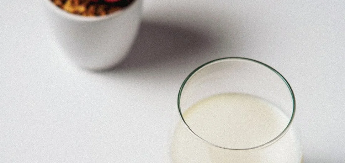 A glass with a pale liquid sits on a clean white surface, with a small potted plant blurred in the background.