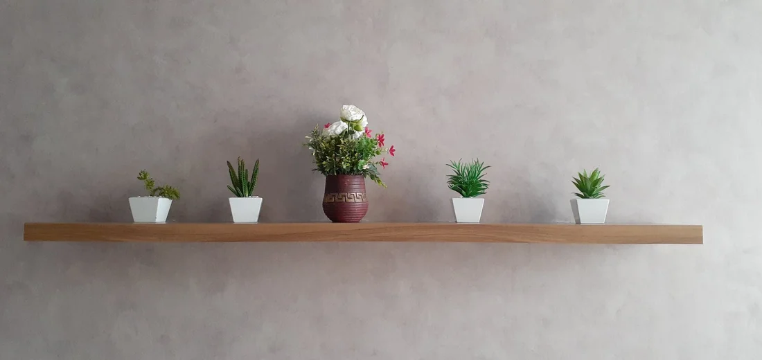 A minimalist wooden shelf mounted on a gray wall displays four small white pots with various succulents, plus a larger brown vase with pink and white flowers in the center.