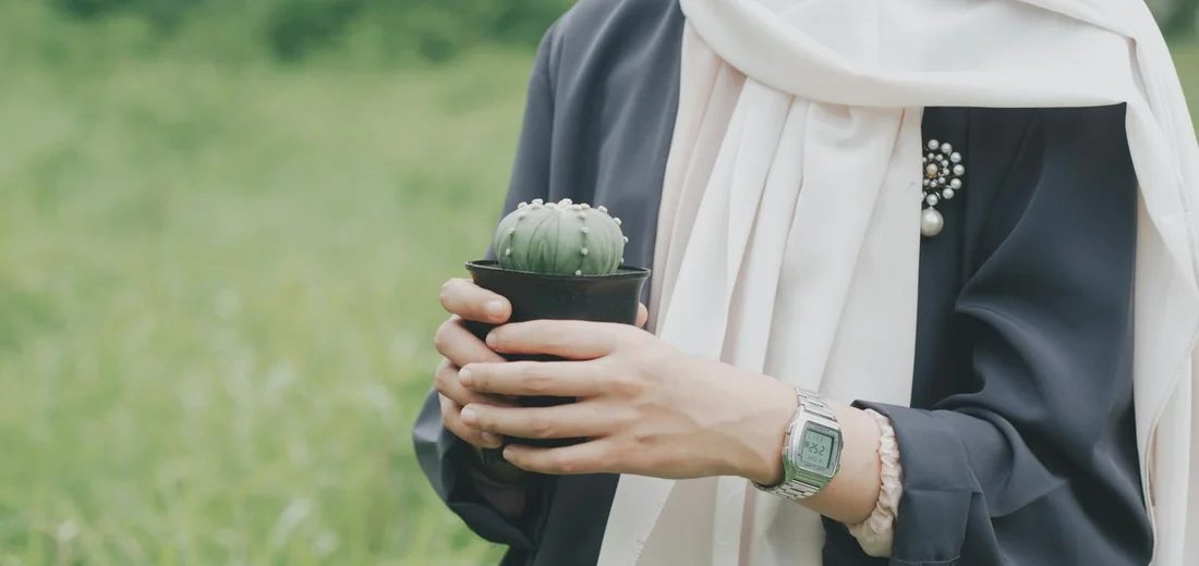 Person wearing a scarf and watch holding a small potted succulent outdoors.