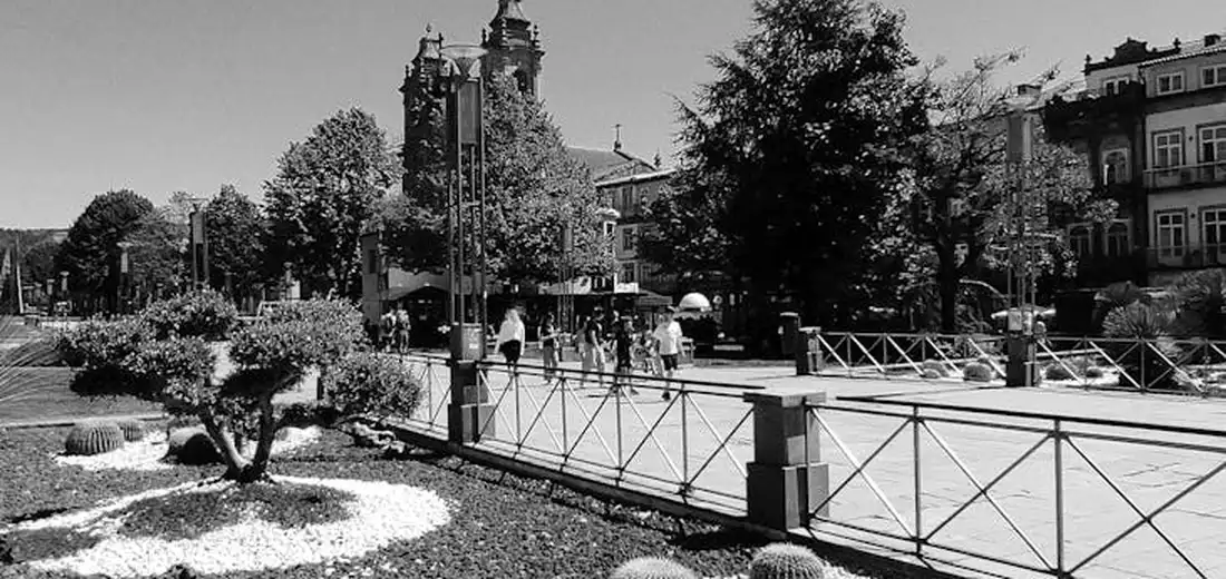 Black-and-white photograph of an urban plaza with a decorative railing, manicured shrubs, and people walking in the background.