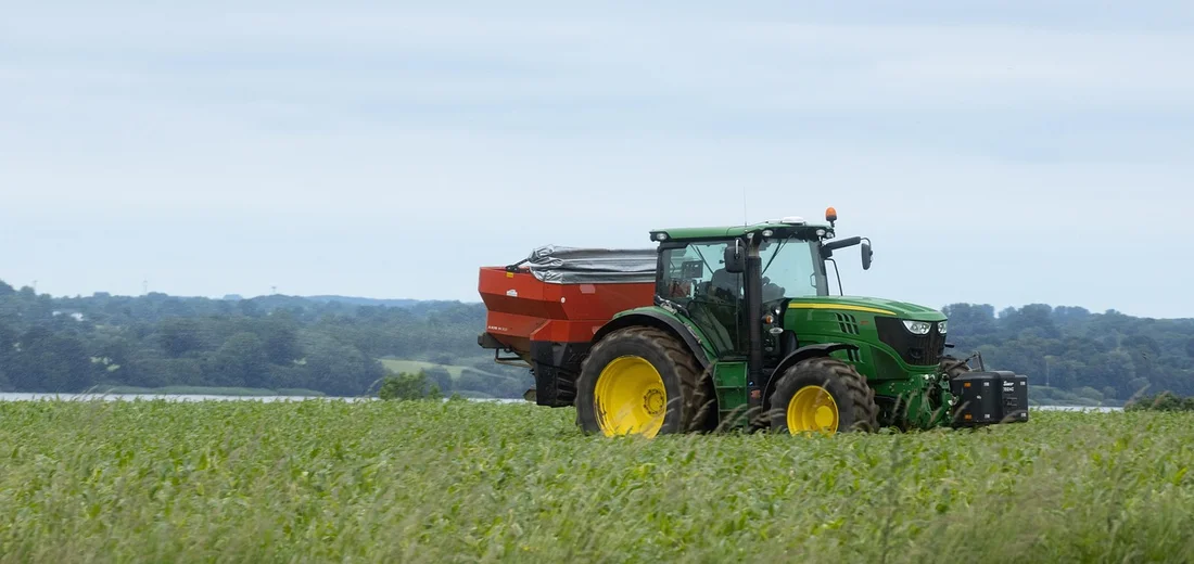 Green John Deere tractor with an orange fertilizer spreader attached, standing in a grassy field under a clear blue sky.