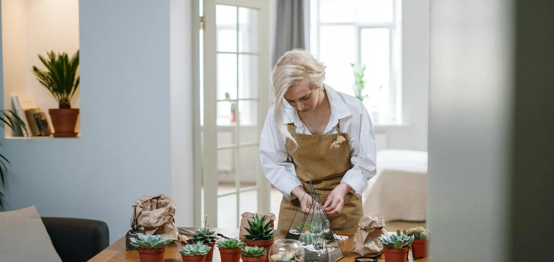 A person wearing a brown apron tends glass terrariums with succulents on a wooden table in a bright, airy room.