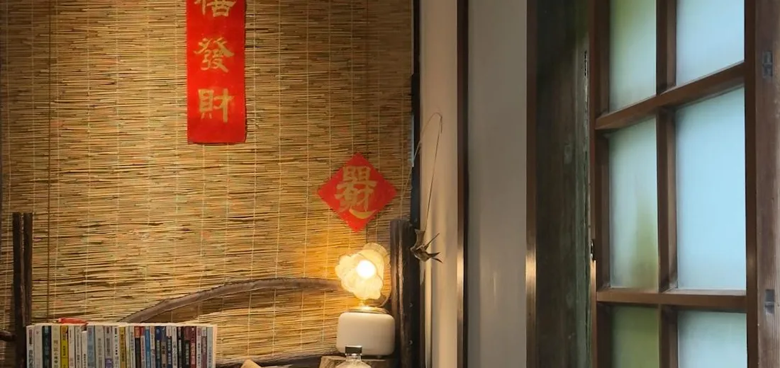 Cozy indoor room with a woven bamboo wall, hanging red banners, a glowing lamp, a small stack of books, and a multi-pane window—used as a backdrop in the ZZ plant care guide.
